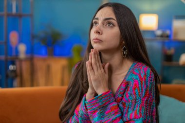 Lütfen, Tanrım yardım et. Genç bayan kollarını kavuşturarak dua ediyor, yukarı bakıyor ve dilek tutuyor, umutlu bir ifadeyle rica ediyor, özür diliyor. Evdeki kanepede oturan üzgün kız.