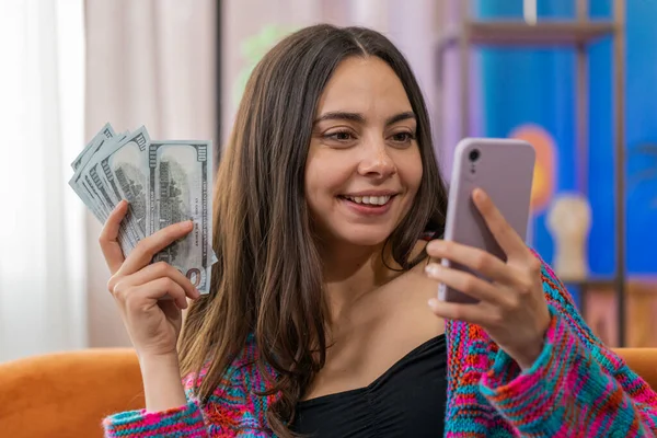 家族の予算を計画してる お金の現金を数える女性は スマートフォンを使用して国内請求書を計算する 楽しい女の子は収入に満足し 計画された休暇のためのお金を節約します ソファの上の家のアパートの部屋で贈り物 — ストック写真
