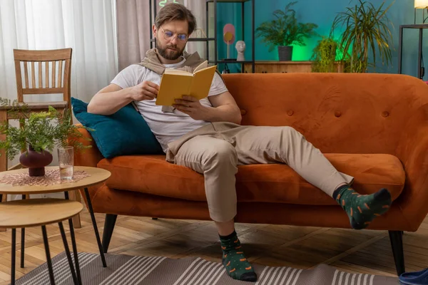 Young man relaxing interesting book, turning pages smiling enjoying literature, taking a rest on comfortable couch. Portrait of peaceful cheerful Caucasian guy at home apartment living room on sofa