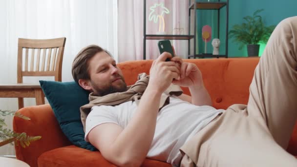 躺在沙发上的年轻人在家里的起居室里用手机微笑 白种人在智能手机社交媒体应用程序上发短信 分享内容 观看轻松电影 — 图库视频影像