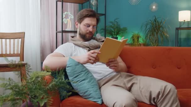 年轻人放松有趣的书 翻开书页 笑着欣赏文学 在舒适的沙发上休息 安详快乐的白人男子在沙发上的公寓客厅的画像 — 图库视频影像