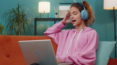 Kulaklıklı beyaz liseli kız dizüstü bilgisayar kullanıyor müzik dinliyor ya da ders dinliyor, uzaktan öğreniyor. Online eğitim. Küçük bir çocuk, çocuk dinleniyor, iş yerinde dinleniyor, kanepede oturuyor.