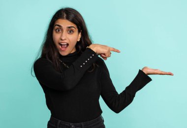 Genç Hintli kadın başparmağını kaldırıyor ve boş bir yeri işaret ediyor. Ticari metinler için reklam alanı, ürün tanıtımı için kopyalama alanı. Arap Hindu kız mavi stüdyo arka planında tek başına kapalı.