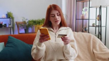 Genç kızıl saçlı bir kadın kredi kartı ve akıllı telefon kullanarak para aktarıyor, internetten nakit para almıyor, ev içinde yemek siparişi veriyor. Mutlu kız kanepede oturuyor.