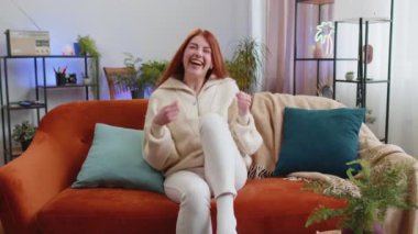 Mutlu kızıl saçlı kadın bağırıyor, başarıyı kutluyor, kazanıyor, gol başarısı iyi haber yumruk tokuşturuyorum modern evde piyango şansı. Oturma odasında kanepede oturan kız.