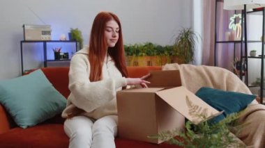 Evdeki kanepede oturan mutlu, genç bir kadın teslimat paketini açıyor. Gülümseyen tatmin olmuş kız alışverişci internet müşterisi karton kutu açıyor hızlı posta yoluyla satın alma hediyesi alıyor