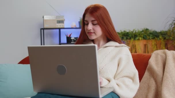自宅のソファでリビングルームで仕事を終えた後 ソファの上に座っている若い女性 オンラインリモートの仕事で働いている赤毛の女の子のフリーランス 電子学習 ノートPc上でインターネットを閲覧 — ストック動画