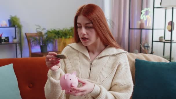 可怜的红头发女人没有足够的钱 在家里的公寓里拿着储蓄罐和一块钱的钞票 金融危机 贫穷和匮乏 坐在沙发上的客厅里的女孩 — 图库视频影像