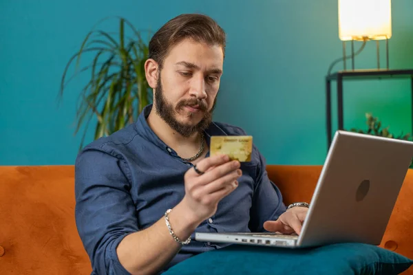 Young lebanese man using credit bank card and laptop computer while transferring money, purchases online shopping, order food delivery. Portrait of arabian guy at home apartment living room on couch