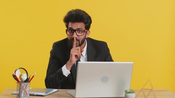 静かにしてください インドのビジネスマンは唇に人差し指を押すと 沈黙ジェスチャーのサインはオフィスの職場の机で秘密を伝えません ヒンズー教徒の男は黄色の背景に隔離されたラップトップで作業 — ストック動画