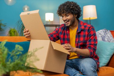 Happy indian man unpacking delivery parcel sitting at home apartment. Smiling satisfied hindu guy shopper, online shop customer opening cardboard box receiving purchase gift by fast postal shipping