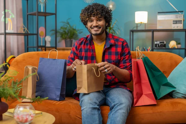Young indian man happy shopaholic consumer came back home after shopping sale with bags. Portrait of hindu guy satisfied received parcels purchase from online order at home apartment room on sofa
