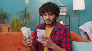 Planning family budget. Smiling indian man counting money cash, use smartphone calculate domestic bills at home room. Joyful hindu guy satisfied of income and saves money for planned vacation, gifts