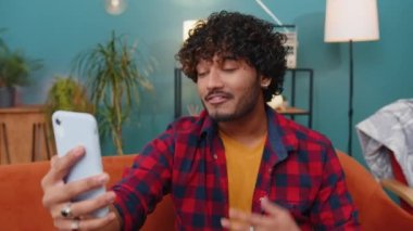 Mutlu Hintli blog yazarı akıllı telefondan selfie çekiyor, sosyal medya takipçileriyle çevrimiçi video görüşmeleri yapıyor, vlog hikayeleri kaydediyor. Evdeki kanepede oturan Hintli genç adam.