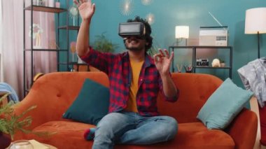 Indian man using virtual reality futuristic technology VR app headset helmet to play simulation 3D 360 video game, watching film movie at modern home apartment. Hindu guy in goggles sitting on couch