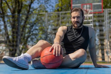 Basketbol maçından sonra spor giyim kuşamında yorgun Lübnanlı adamın portresi. Şehir parkının kortunda rahatça oturup topu tutan adam. Spor sahasında spor rutini. Motivasyon. Açık havada.