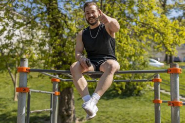 Spor giyim sektöründeki atletik adam olumlu tepkiler veriyor. Çocuk parkındaki adam yatay barda oturuyor. Spor sağlık, spor rutini, egzersiz. Güç ve motivasyon. Açık spor salonu.