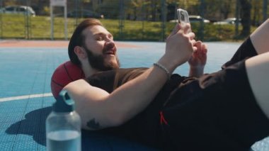 Basketbol maçından sonra spor giyim kuşamında yorgun Lübnanlı adamın portresi. Top rahatlatıcı bir şekilde uzanan adam şehir kortunda mesajlaşarak akıllı telefon kullanıyor. Spor sahasında spor