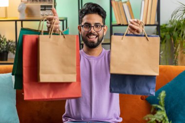 Genç Hintli adam alışveriş kolik tüketicisi çantalarla birlikte eve döndü. Hintli adamın portresi, evdeki kanepede internetten alınmış paketler aldı.