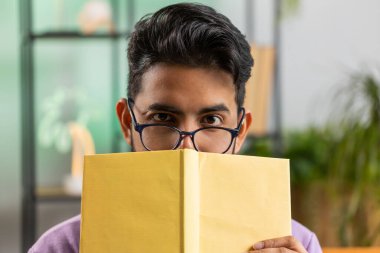 Gözlüklü genç Hintli adam ilginç kitaplar okuyor, kitapların arkasına saklanıyor, edebiyattan zevk alıyor, kanepede dinleniyor. Evdeki kanepede oturan mutlu Hintli adamın portresi.