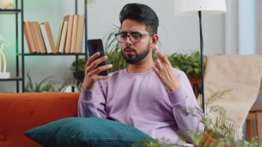 Sad displeased indian man use smartphone typing browsing, loses becoming surprised sudden lottery results, bad news, fortune loss, fail, deadline, virus. Young hindu guy at home living room on couch
