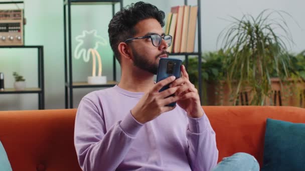 Indian Man Sitting Sofa Uses Mobile Phone Smiles Home Living — Vídeos de Stock