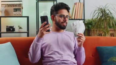 Aile bütçesi planlamak. Gülümseyen Hintli adam nakit para sayıyor, akıllı telefon kullanıyor ev faturalarını hesaplıyor. Neşeli Hintli adam gelirinden memnun ve planlı tatil ve hediyeler için para biriktiriyor.