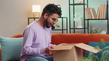 Happy indian man unpacking delivery parcel sitting at home apartment. Smiling satisfied hindu guy shopper, online shop customer opening cardboard box receiving purchase gift by fast postal shipping