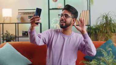 Mutlu Hintli blog yazarı akıllı telefondan selfie çekiyor, sosyal medya takipçileriyle çevrimiçi video görüşmeleri yapıyor, vlog hikayeleri kaydediyor. Evdeki kanepede oturan Hintli genç adam.