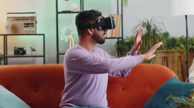Indian man using virtual reality futuristic technology VR app headset helmet to play simulation 3D 360 video game, watching film movie at modern home apartment. Hindu guy in goggles sitting on couch