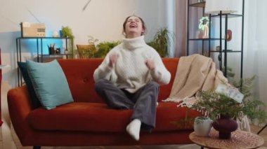 Money cash dollar rain falling on woman celebrating success, winning, goal achievement, good news, lottery jackpot luck, dream come true at home apartment indoors. Career grow. Girl sits on sofa