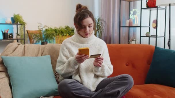 妇女在汇款时使用信用卡和智能手机 在网上购物时没有现金 在家里的公寓里订购食物 快乐的小女孩坐在沙发上的房间里 — 图库视频影像