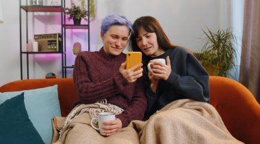 Two young and cheerful lesbian or friends hold their smartphones while shopping on e-commerce apps at home living room. Girls enjoy having fun browsing different social media applications together