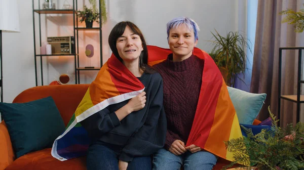 Two lesbian women family couple or girl friends holding LGBT people gay pride flag in hands at home living room. Gay, bisexual, transgender social movements. Concept of happiness freedom love same-sex