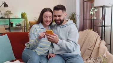 Cep telefonu kullanan aile erkeği kadın, büyük ikramiyeyi kutlayan iyi haberleri buldu. Şanslı piyango zaferini, evde online oyun alışverişi yaparak kutluyor. Karı koca kanepede yan yana.