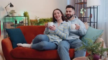 Genç, gülümseyen bir aile evli bir erkek kadın sabah evde sıcak kahve ya da bitki çayı içerek turuncu kanepede yatıyor. Karı koca birlikte rahatça dinleniyor.