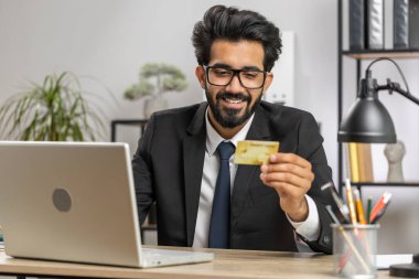 Sakallı iş adamı yazılım geliştirici kredi kartı ile online satın alma alışverişi yapıyor ve ofis masasında dizüstü bilgisayar kullanıyor. Hintli serbest çalışan iş adamı