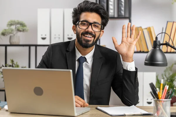 stock image Hello. Indian business man working on laptop computer smiling friendly at camera and waving hands gesturing hi, greeting or goodbye, welcoming with hospitable expression at home office workplace desk