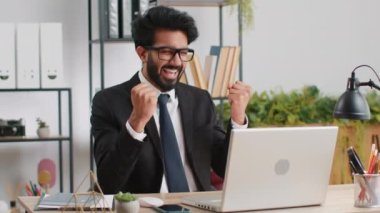 Indian businessman fingers crossed working on laptop at home office desk typing browsing say Wow yes found out great big lottery jackpot win, good job news, celebrating. Manager freelancer arab man