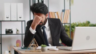 Tired ill indian businessman suffering from headache problem tension and migraine, stress at home office desk workplace. Bearded freelancer broker worker man works on laptop notebook. Overworking