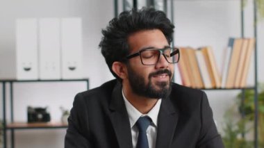 Modern ofis çalışma masasındaki mutlu, sakallı Hintli işadamının portresi başını kameraya çevir. Resmi takım elbiseli bir erkek Hindu. Yakışıklı Uzaktan kumandalı adam dizüstü bilgisayarla çalışıyor