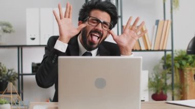 Indian businessman hiding behind laptop computer, making funny face, fooling around, putting his tongue out remote working disrespecting someone at office desk workplace. Professional freelancer man