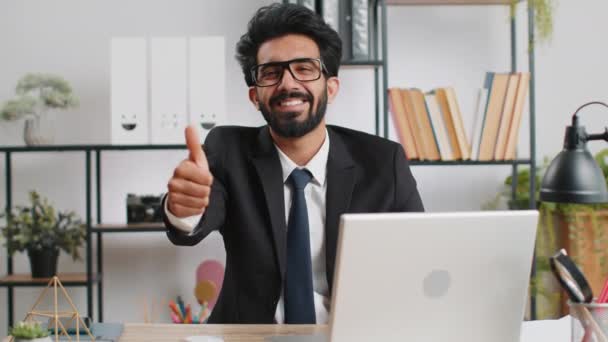 まるで 幸せな若いインドのビジネスマンの男は オフィスの職場で肯定的な何か良い 肯定的なフィードバックにサインアップなど 親指を表示するカメラで承認的に見てラップトップに取り組んでいます フリーランスの男 — ストック動画