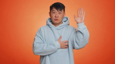 Dürüst olacağıma yemin ederim. Saygıdeğer Asyalı yakışıklı adam yemin etmek için elini kaldırıyor, dürüst olacağına ve doğruyu söyleyeceğine söz veriyor, elini göğsüne koyuyor. Yetişkin adam turuncu stüdyoda izole edilmiş.