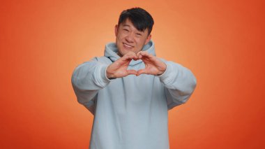 Man in love. Smiling asian man 40 years old makes heart gesture demonstrates love sign expresses good feelings and sympathy. Handsome chinese adult guy isolated alone on orange studio background