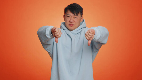 Не нравится. Расстроенный недовольный азиат в толстовке с опущенными большими пальцами знак жеста, выражая недовольство, неодобрение, недовольство, неприязнь. Красивый взрослый парень. Крытая студия на оранжевом фоне