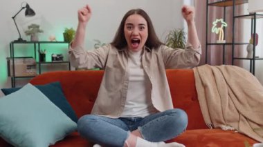 Happy young woman shouting, celebrating success, winning, goal achievement good news raising fists in gesture I did it, lottery luck at modern home apartment. Girl in living room sitting on couch