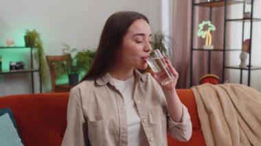Susamış genç bir kadın elinde bir bardak su tutarak susuzluğu gidermek için bir yudum su içiriyor. Oturma odasında oturuyor. İyi yaşam alışkanlıkları, sağlıklı zayıflama, kilo verme konsepti olan bir kız.