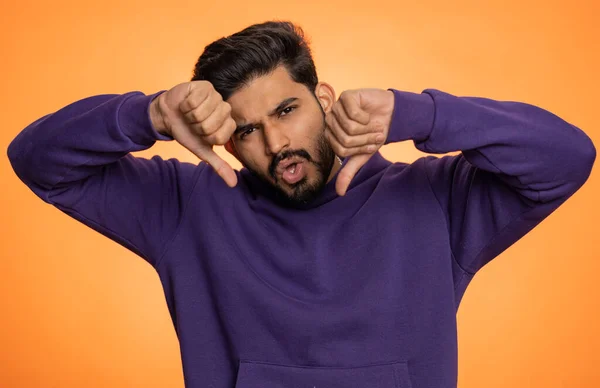 Dislike. Upset unhappy indian man in hoodie showing thumbs down sign gesture, expressing discontent, disapproval, dissatisfied, dislike. Handsome young guy. Indoor studio shot on orange background