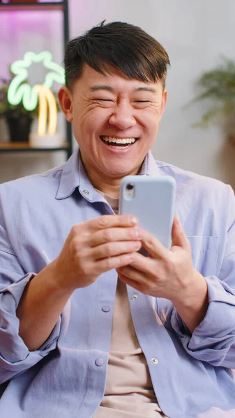 幸せなアジアの勝者の男性は 携帯電話の入力のブラウジングを使用すると言うすごいはい大きな勝利良いニュースを発見宝くじのジャックポットを祝う ソファに座っている部屋で自宅で中国の大人の男 — ストック写真